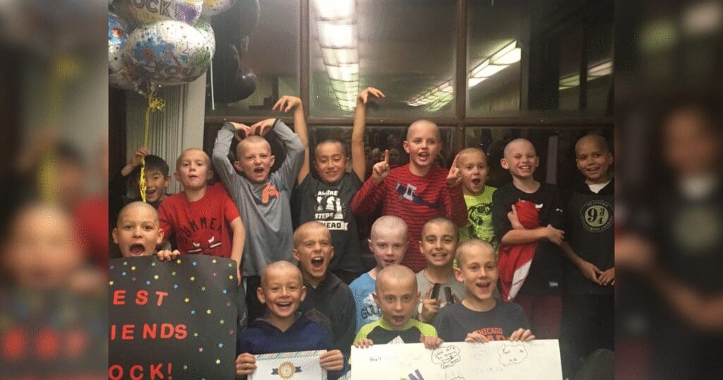 A group of smiling children with shaved heads pose excitedly while holding supportive signs and balloons in a brightly lit room. The mood is joyful and triumphant.