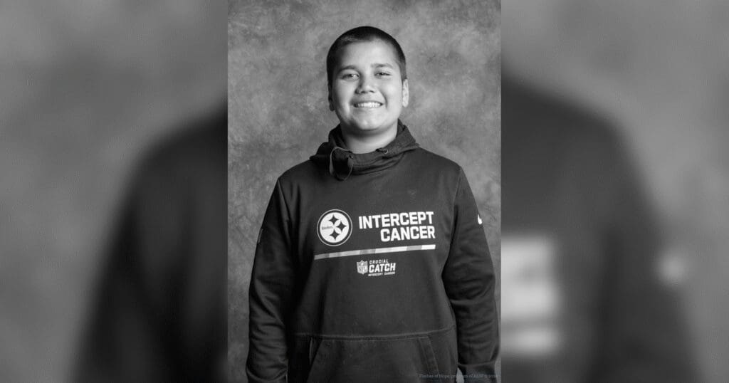A smiling boy stands confidently against a subtle textured background, wearing a hoodie with "INTERCEPT CANCER" text, exuding positivity and strength.