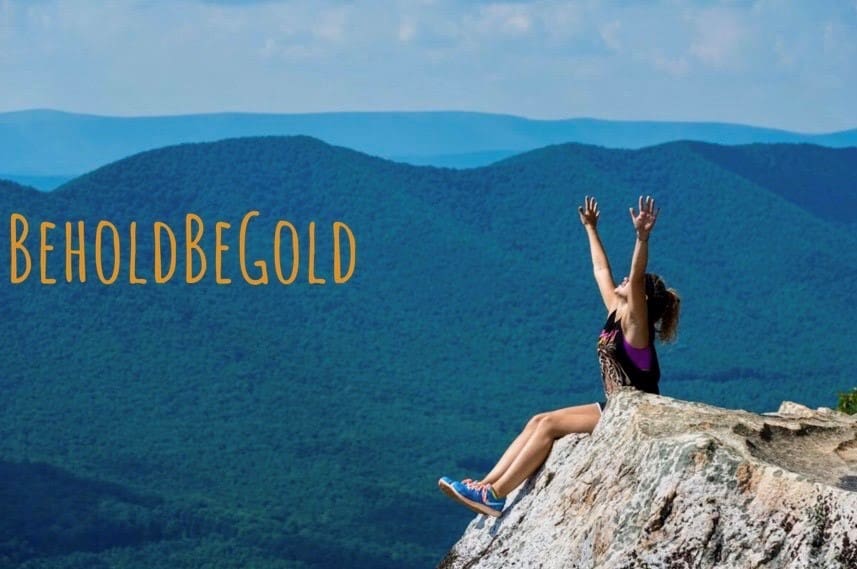 A person sits triumphantly on a rock ledge with arms raised against a vast, mountainous backdrop. The text "BeholdBeGold" appears on the left.