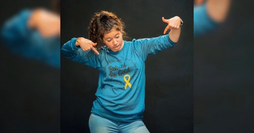 A woman in a blue shirt with a yellow childhood cancer awareness ribbon points at a message on her shirt, expressing confidence. The background is dark, highlighting her pose and expression.
