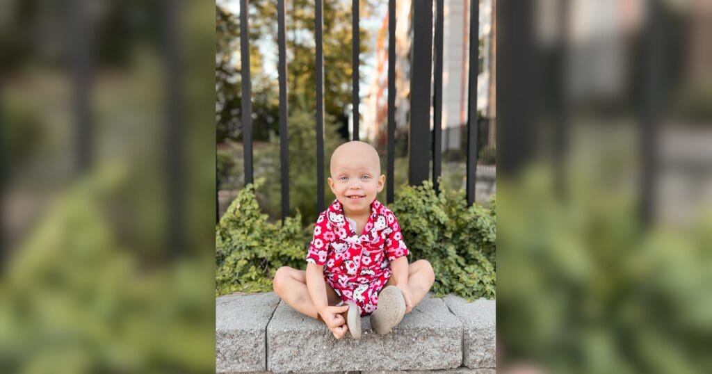 A smiling child in a bright red floral outfit sits cross-legged on a stone ledge. Green foliage and a black fence form the backdrop, creating a cheerful scene.