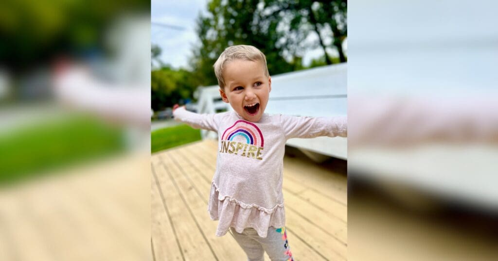 A joyful child with short hair and a wide smile stands on a wooden deck, arms outstretched. They wear a shirt with a rainbow and "INSPIRE" text. Trees are in the background.