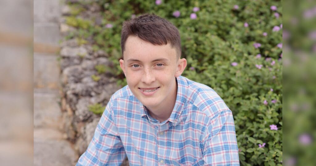 Teen boy in a plaid shirt smiles warmly, seated outdoors. Background features lush greenery with small purple flowers, creating a serene vibe.