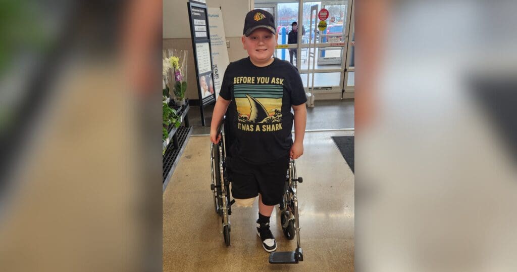 A boy wearing a baseball cap and a shirt with a shark print stands smiling beside a wheelchair in a store entrance, conveying positivity and resilience.