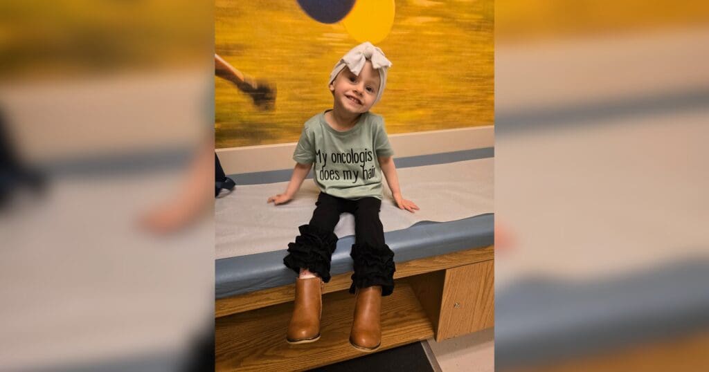 A smiling child with a headband sits on a medical examination table, wearing a T-shirt saying "My oncologist does my hair." The mood is uplifting.