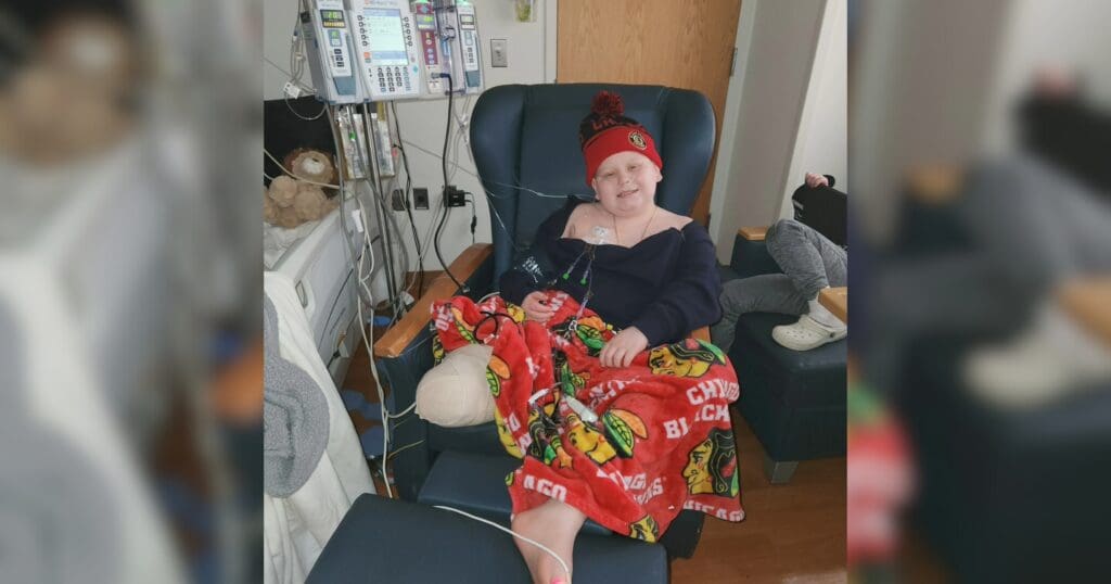 A child in a hospital room sits in a chair, smiling warmly. They wear a beanie and are wrapped in a colorful sports team blanket, conveying cheerfulness and comfort.