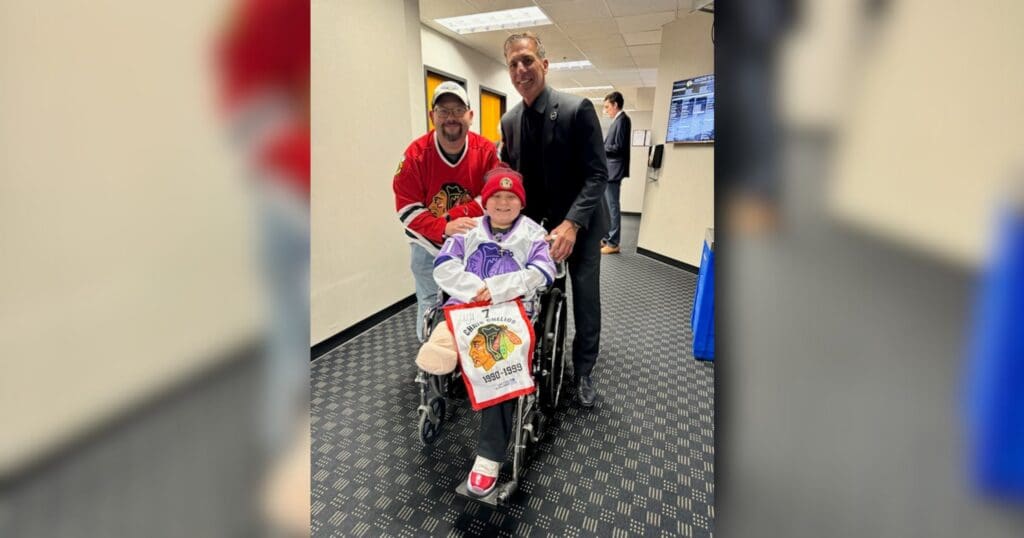 A smiling child in a wheelchair, flanked by two individuals. One wears a red hockey jersey, the other a black suit. They are in a carpeted hallway.