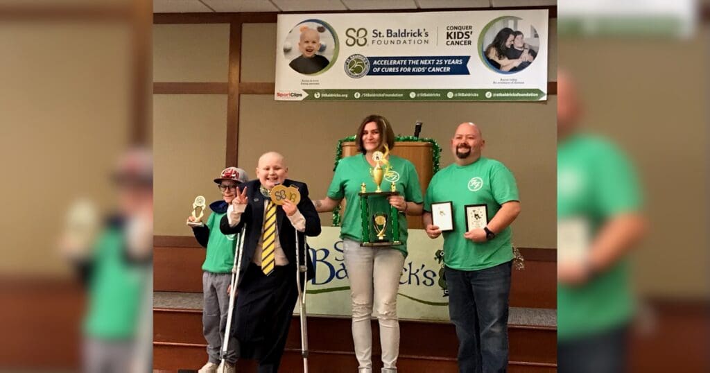 Four people on stage celebrate at a St. Baldrick's Foundation event, holding trophies and plaques. The mood is joyful and triumphant.