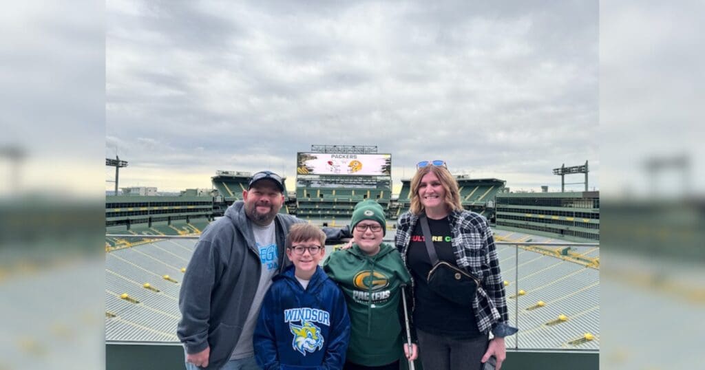 A smiling family of four stands in a stadium with a large screen and empty seating behind them. They're dressed warmly, suggesting a cool day.