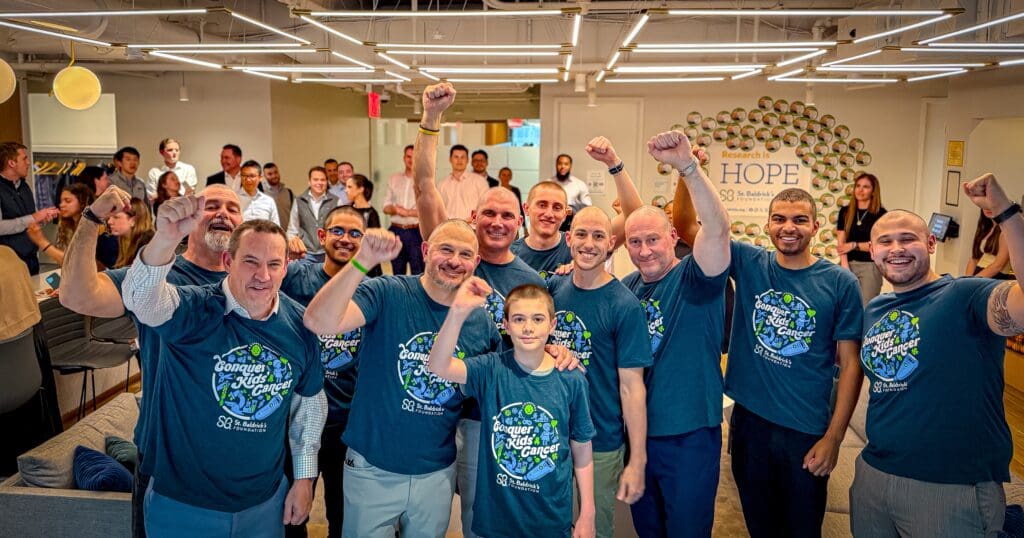 A group of smiling people wearing matching "Conquer Kids' Cancer" t-shirts raise their fists in a bright room, conveying a sense of unity and hope.