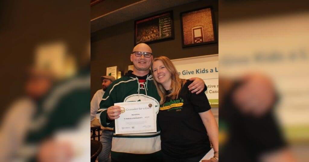A smiling man in a sports jersey holds a certificate, standing beside a woman in a black shirt. They pose at an indoor event, conveying a joyful atmosphere.