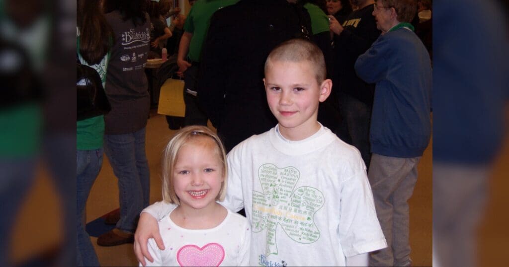 Two kids smiling; a girl in a white shirt with a pink heart and a boy in a white T-shirt with a green graphic. Crowd in the background, casual setting.