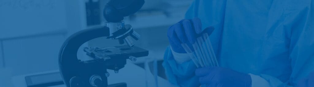 A person in blue lab attire and gloves holds test tubes near a microscope in a laboratory, indicating a scientific research setting.