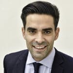 Smiling man in a suit with dark hair, set against a plain background. He exudes confidence and professionalism, creating a positive and approachable tone.