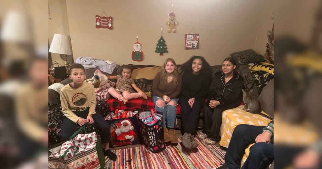 A group of five children sit on a couch, surrounded by Christmas decor and gifts. The room is warmly lit, creating a cozy holiday atmosphere.