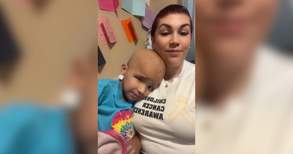 A woman and child are sitting together, with the child resting on the woman's chest. The woman wears a "Childhood Cancer Awareness" shirt. Colorful notes are on the wall behind them, creating a supportive atmosphere.