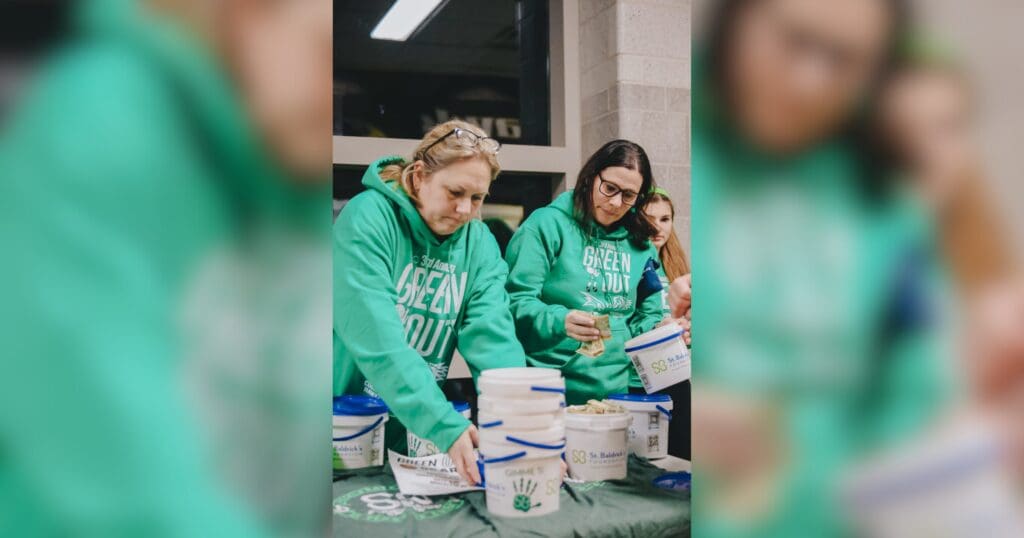 Two women in green hoodies are engaged in a fundraising event, handling donation buckets on a table. Their focused expressions convey dedication.