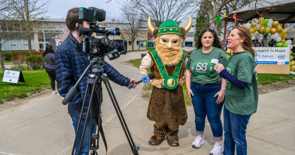 A person in a Viking costume stands between two people being interviewed outdoors. A cameraman records the scene. The mood is lively and festive.