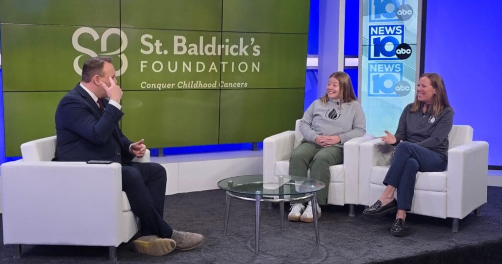 A news anchor interviews two women on a TV set. Behind them, the St. Baldrick's Foundation logo and a slogan about conquering childhood cancers are displayed.