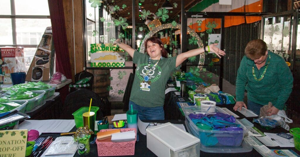 A joyful woman in a green St. Patrick's Day-themed room spreads her arms wide, smiling. Decorations and donation items surround her. Another person is sorting papers.