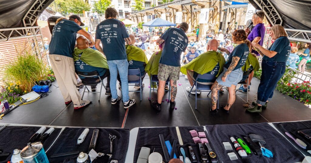 Barbers on an outdoor stage shave heads of seated individuals. A crowd observes the event. Tools are organized in the foreground. The scene is lively and communal.