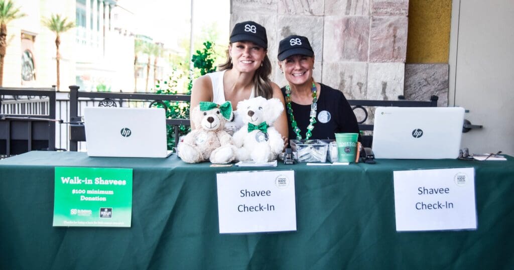 Two smiling individuals in matching black hats sit behind a check-in table with laptops, teddy bears, and donation signs for a charity event.