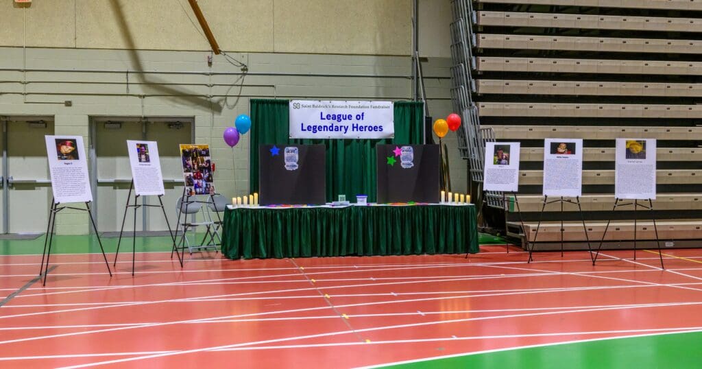 A display titled "League of Legendary Heroes" is set up in a gym. It features a green table with lit candles, colorful balloons, and informational posters on stands. The setting is bright and celebratory.