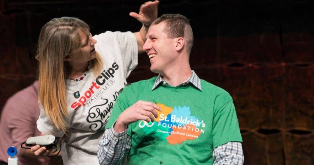 A woman wearing a "Sport Clips" shirt shaves a smiling man's head, who is wearing a "St. Baldrick's Foundation" shirt, promoting cancer awareness.
