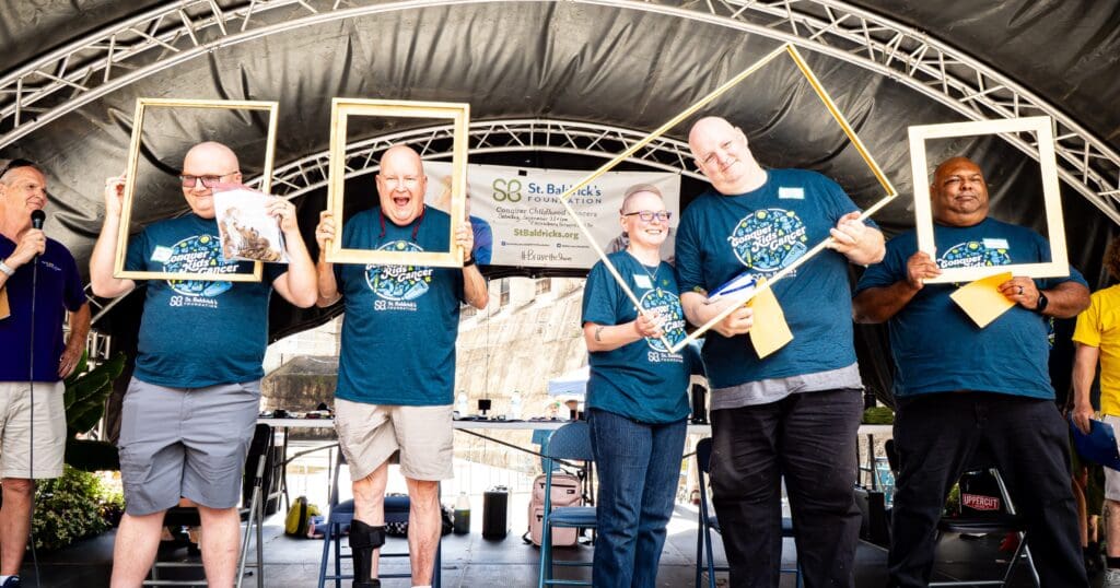 A group of five smiling people on stage hold empty wooden frames. They wear matching blue event T-shirts. The backdrop is an archway with event banners.