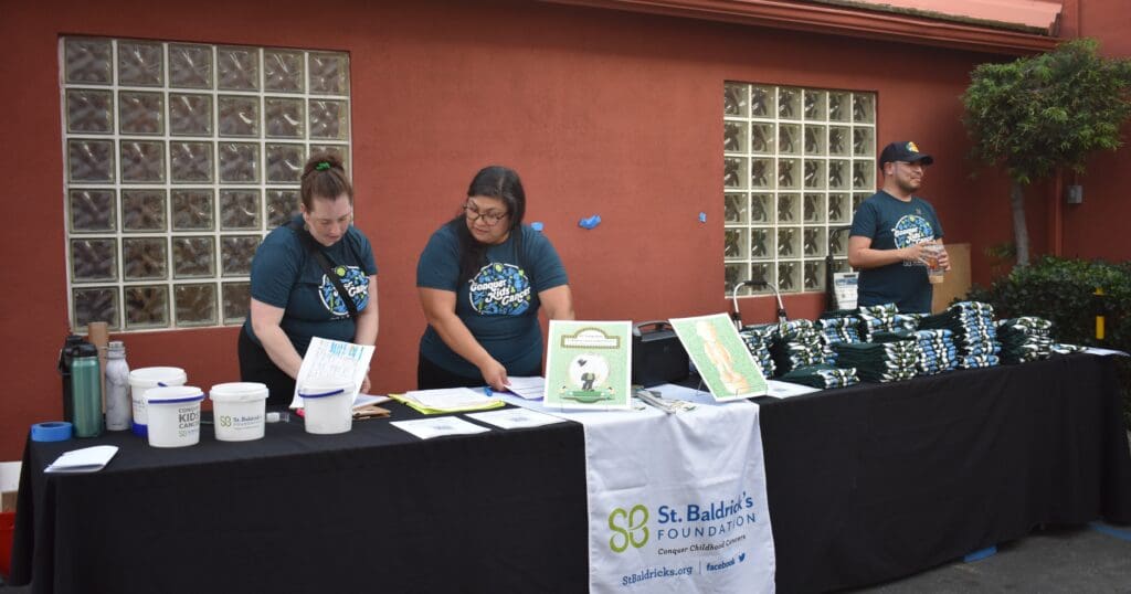 Three people stand behind a table labeled "St. Baldrick's Foundation." The table has donation buckets, papers, and T-shirts. They appear focused and organized.