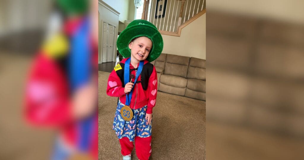A smiling child in a colorful clown outfit, featuring a green hat, red shirt with heart patterns, and blue-patterned trousers, stands in a cozy living room.