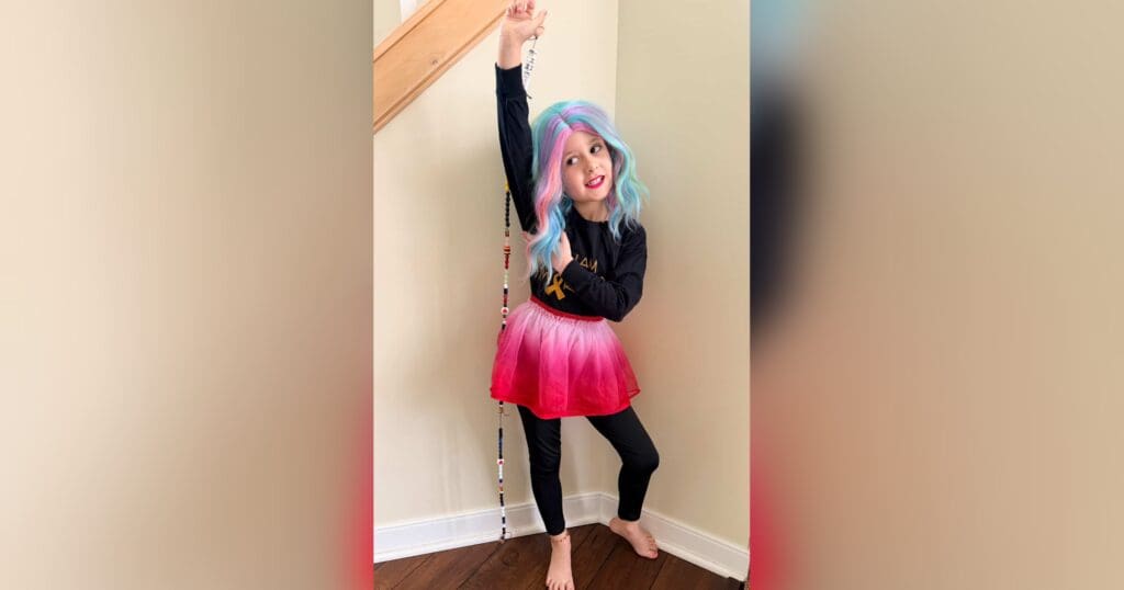 A child in a colorful wig and a pink skirt playfully poses indoors. Her expression is joyful, holding a beaded string beside a staircase railing.
