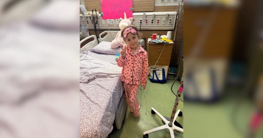 A young child in pink pajamas and a matching headband stands beside a hospital bed with a unicorn stuffed animal on top. A bright sign is visible in the background.