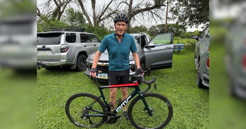 Man in cycling gear stands with a BMC bike in a grassy area, smiling. Parked cars and trees in the background. Casual and outdoor atmosphere.