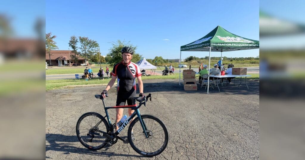 A cyclist in gear poses with a bike in a sunny outdoor setup. People sit under a green tent in the background. The atmosphere is relaxed and casual.