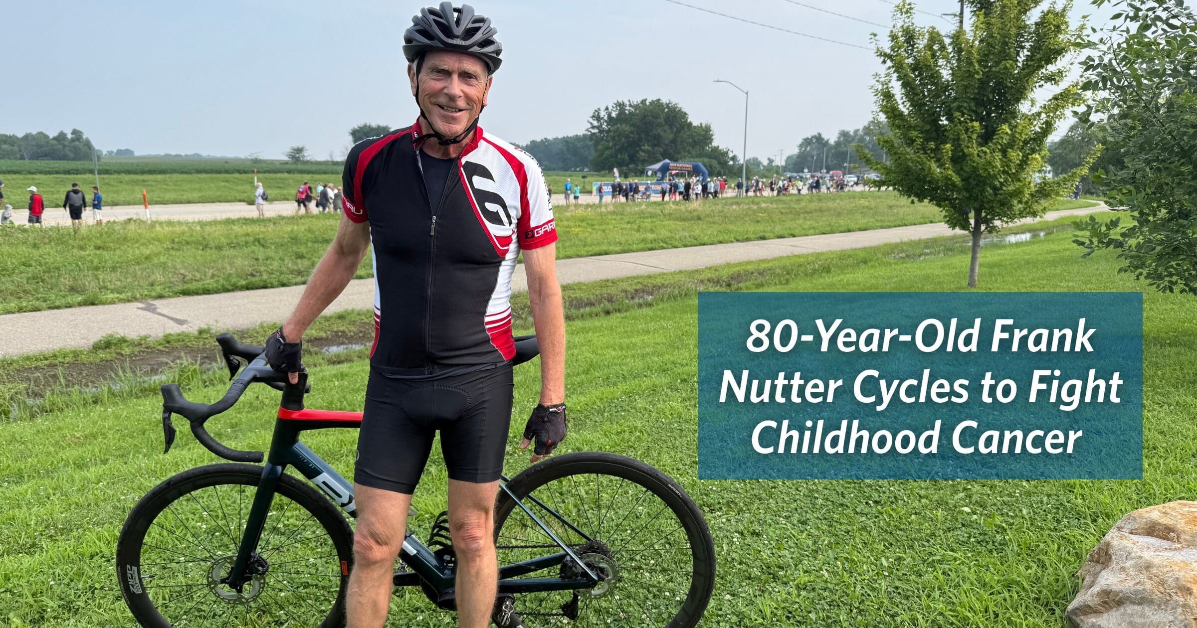An elderly cyclist in black and red gear poses with his bike on a grassy path. A sign reads, "80-Year-Old Frank Nutter Cycles to Fight Childhood Cancer."