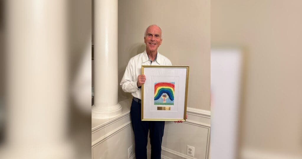 A smiling man in a white shirt holds a framed picture of a colorful rainbow with a gold plaque. The setting is indoors with light-colored walls and a column.