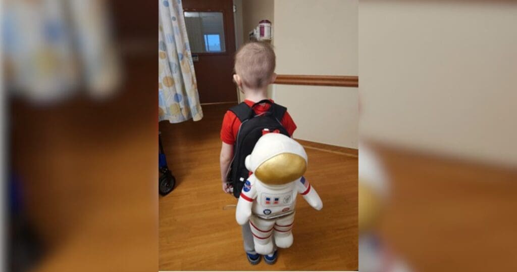 A child in a red shirt stands in a room with wooden floors, wearing a backpack shaped like an astronaut. The scene conveys a sense of adventure.