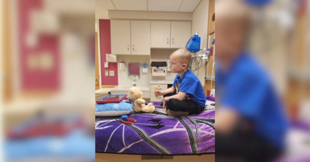 A young child in a blue shirt sits on a hospital bed with a purple blanket, playing with a teddy bear and toy medical kit, creating a comforting scene.