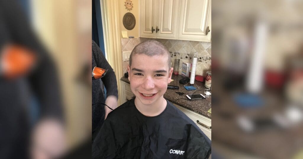 A smiling person with a freshly shaved head sits in a kitchen. He wears a black hairdressing cape. The background shows cabinets and countertop items, conveying a casual and happy atmosphere.