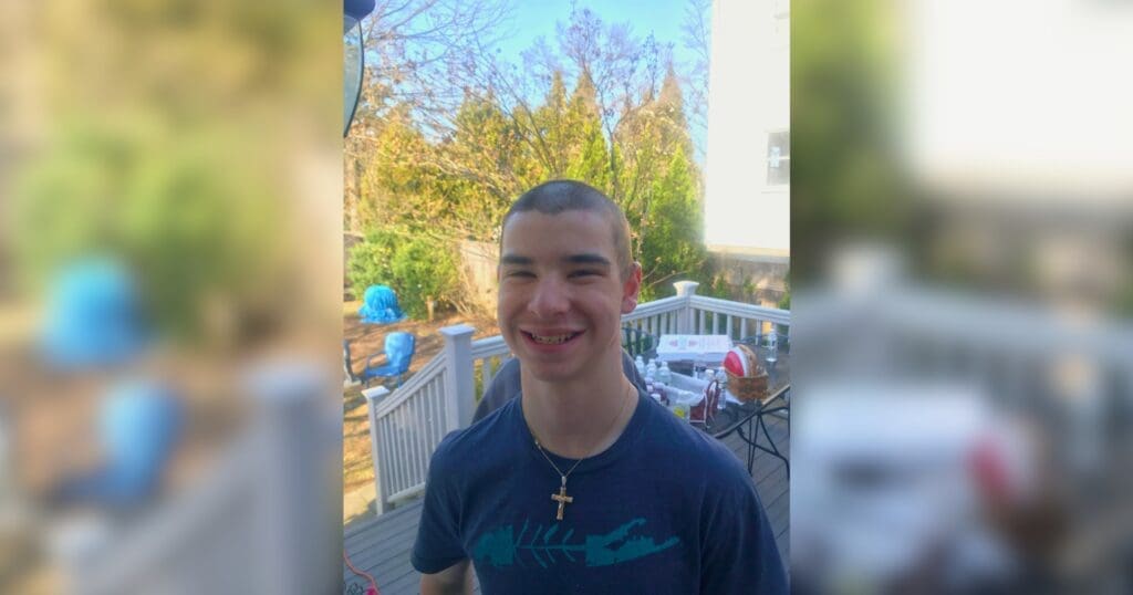 A young man smiles on a sunny patio, wearing a dark shirt and a cross necklace. Outdoor furniture and trees are visible in the background.