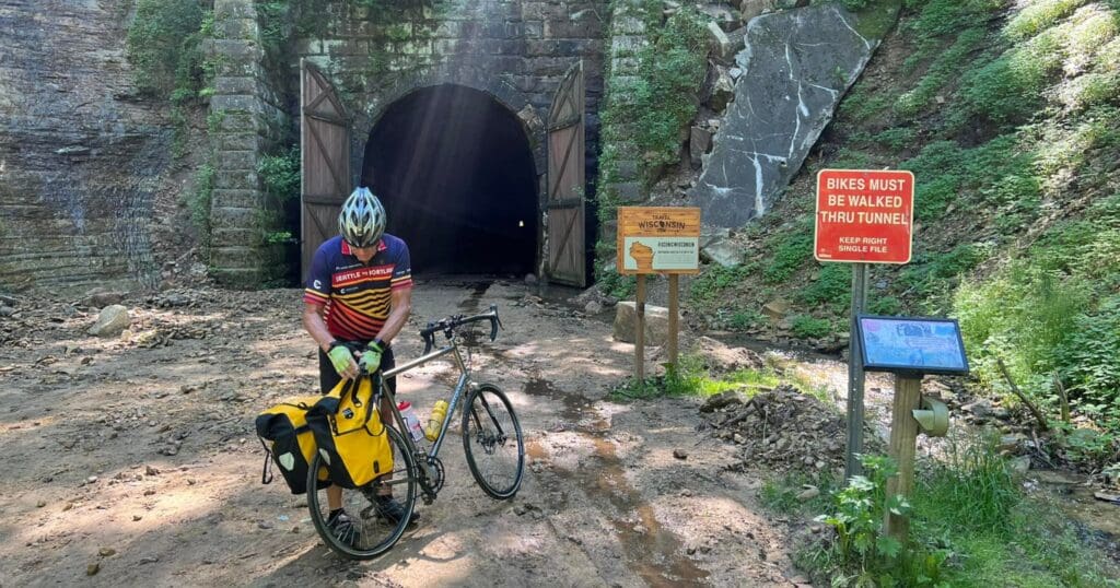Cyclist in colorful gear stands by a bike with yellow panniers near a tunnel entrance surrounded by greenery. Signs warn to walk bikes through. Sunlight beams down.