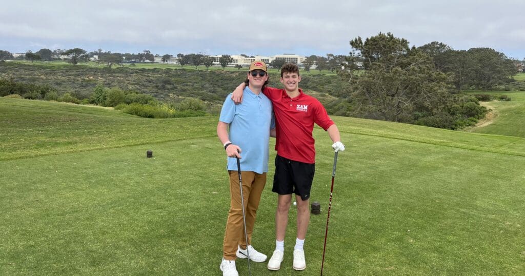 Two men smiling on a golf course, one in a blue shirt and khaki pants, the other in a red shirt and black shorts, each holding a golf club. The background features rolling green hills and a cloudy sky.