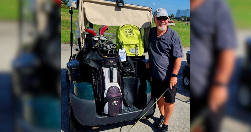 A smiling man stands next to a golf cart holding a golf club. The cart has bags with clubs and a bright bag labeled "Conquer Kids' Cancer." It's a sunny day.