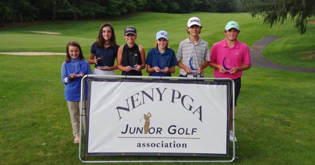Six young golfers stand smiling on a lush golf course, holding trophies. They are behind a sign reading "NENY PGA Junior Golf Association."