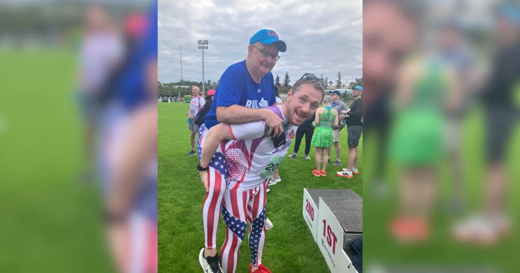 A man in USA flag leggings gives a piggyback ride to another man in a blue shirt and cap. Both smiling, amidst a grassy field with people and a podium.