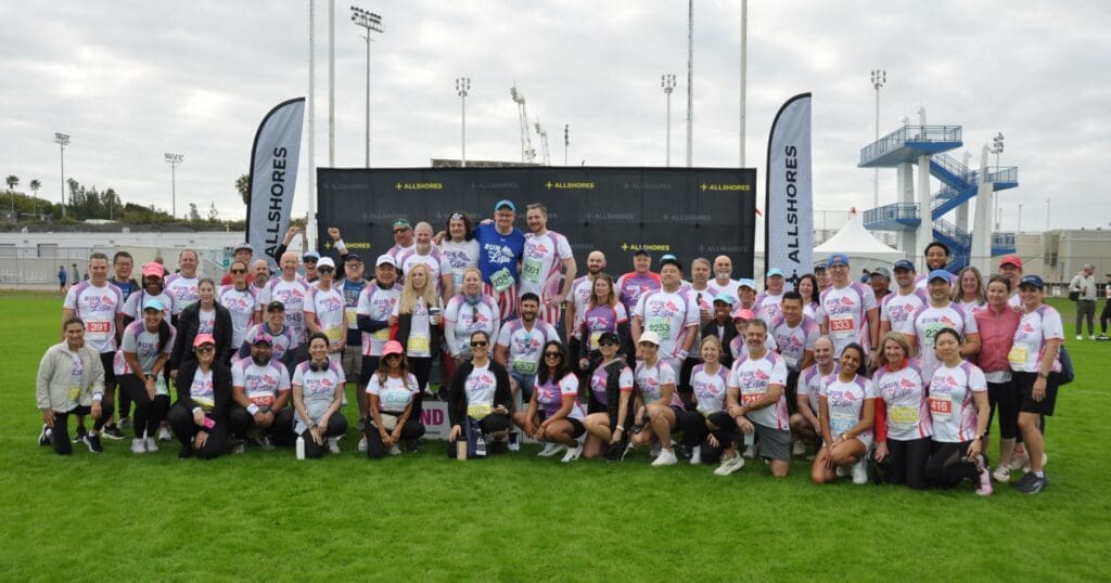 A large group of people in athletic gear pose together on a grassy field for a charity event, "Run for Lisa," supporting kids with cancer.