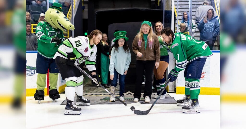 A hockey player in green prepares for a faceoff, flanked by a teammate and a mascot. Two girls, dressed in festive green attire, stand nearby on the ice with smiles, creating a joyful and spirited atmosphere. Spectators watch from the stands.