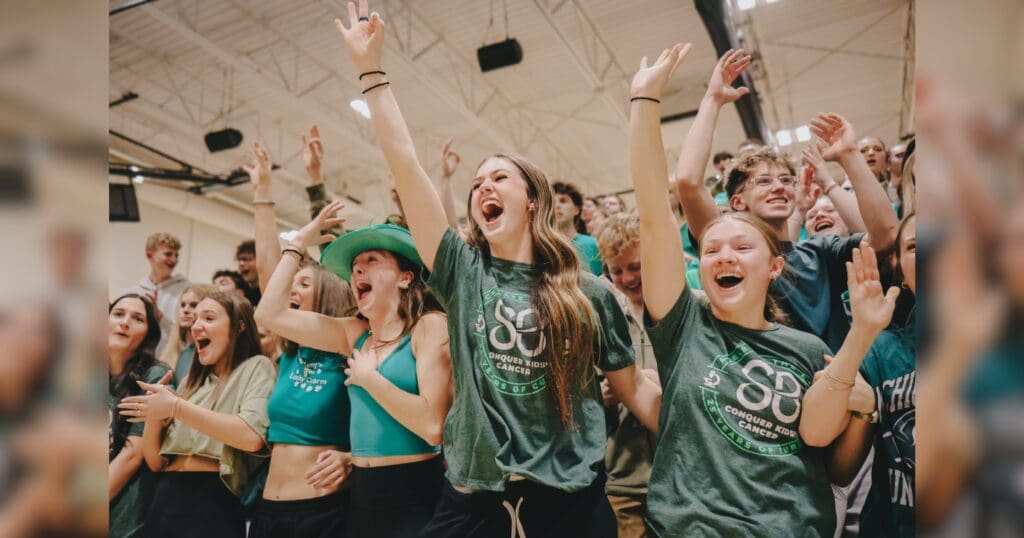 A group of excited students, wearing green shirts, cheer energetically in a gymnasium. Their expressions convey joy and unity during a lively event.