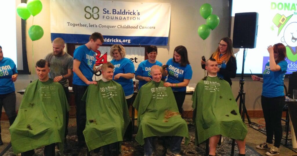 Four people get their heads shaved at a St. Baldrick's Foundation event. Behind them, volunteers in blue shirts smile. Green balloons are visible above.